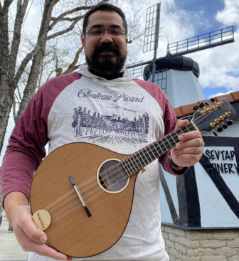 Ian Martyin, with beard and glasses, smiling, holding a mandolin in front of a windmill.