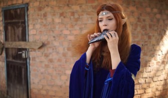 Firamiel, playing ocarina, in front of a brick tower-like building, wearing a blue robe and silver circlet.
