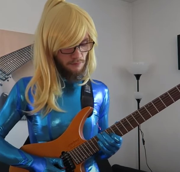 Bad Hairline playing guitar dressed as Zero Suit Samus. He's wearing a blonde wig and a blue tight body suit. He has glasses and a beard.