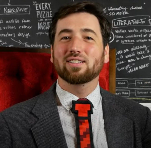 Screenshot of the Games Professor, talking in front of a blackboard with games narrative stuff written in the background. The Prof has brown eyes, short brown hair, and a short beard. He's wearing a suit with a clip on pixelated tie over top a regular red tie. He's also sitting in a red, throne-like chair.